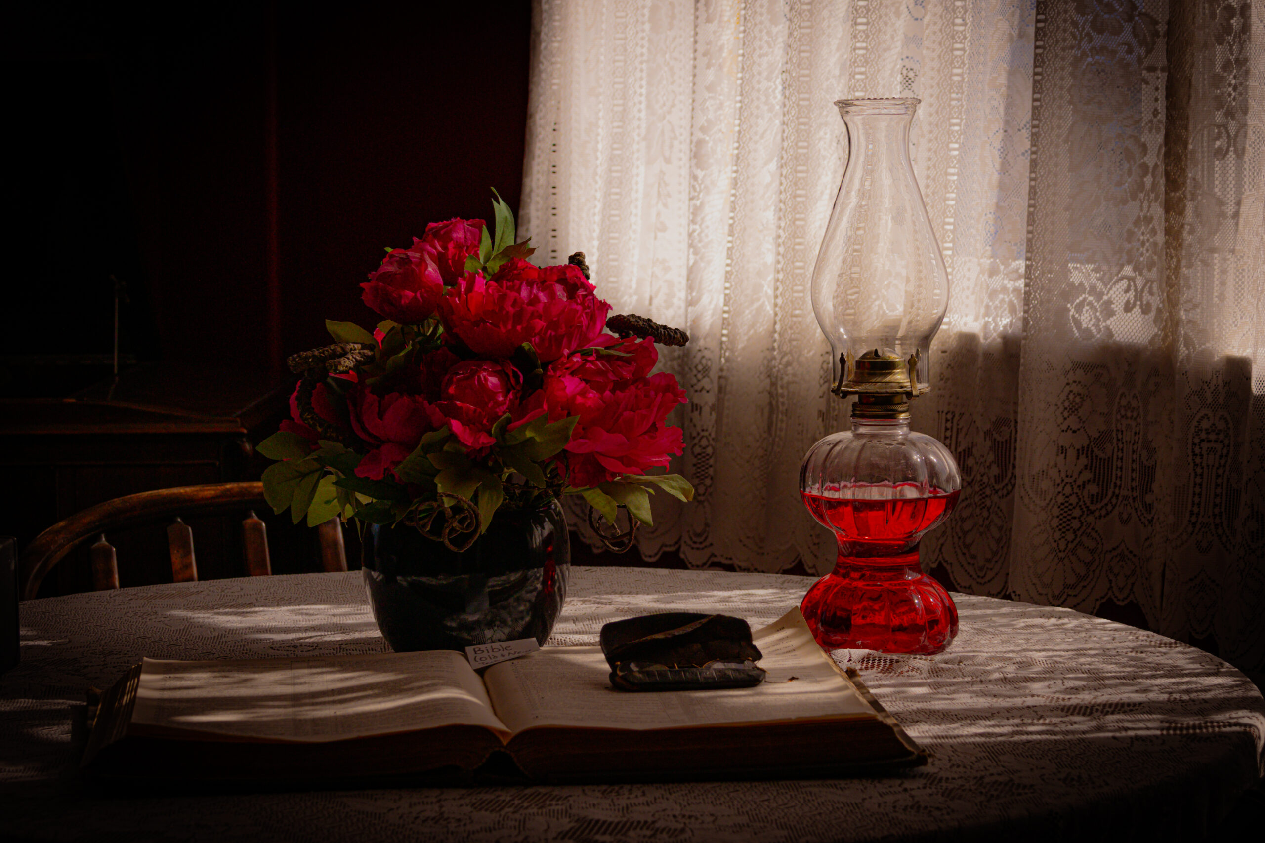 A dark room with lace vurtains over the window shines a dull light over a table. On the table there is a large bible, opened, a glasses case, red flowers in a vase, and an oil lamp with red oil.