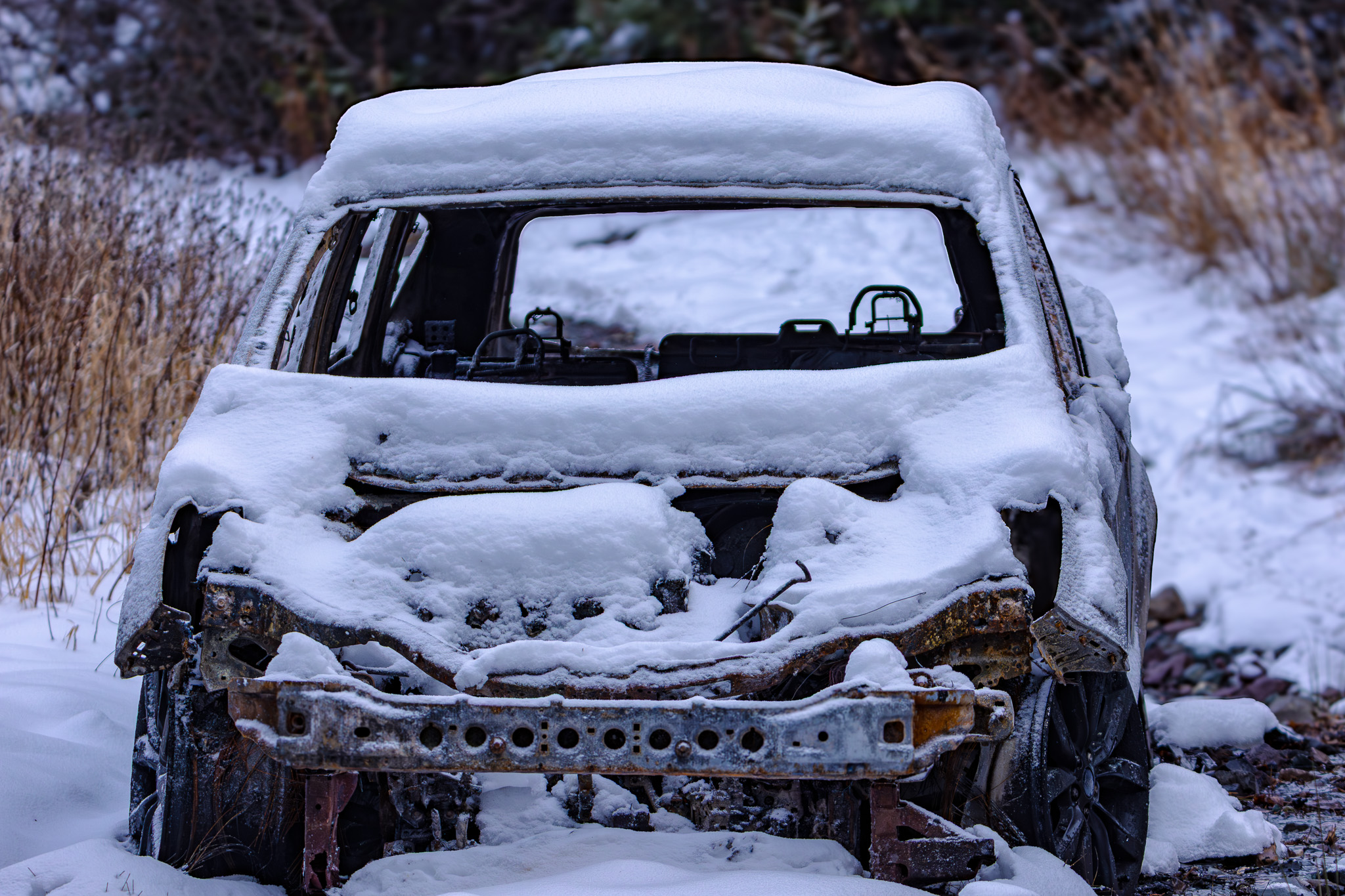 A burnt out car in the middle of a hiking trail, covered in snow. Nothing is left except the metal of the vehicle, having been set on fire.
