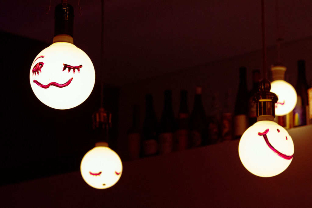 Hanging round lightbulbs with purple painted emoting faces on them. One is winking with a squiggly smile, another is a classic happy face, and another looks to have closed eyes and a solemn expression.
