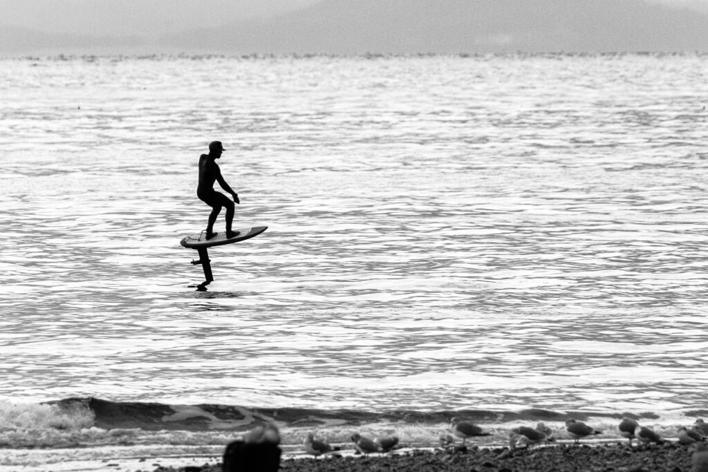 A black and white image of a person in a wet suit on a hoverboard, surfing on calm water in the winter. The surfer is silhouetted against a silvery water background. The hoverboard is quite high out of the water.