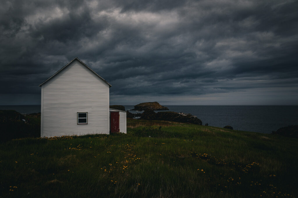 A two-story white house with one small window and a red back door stands alone on a dramatic cliff under moody clouds.