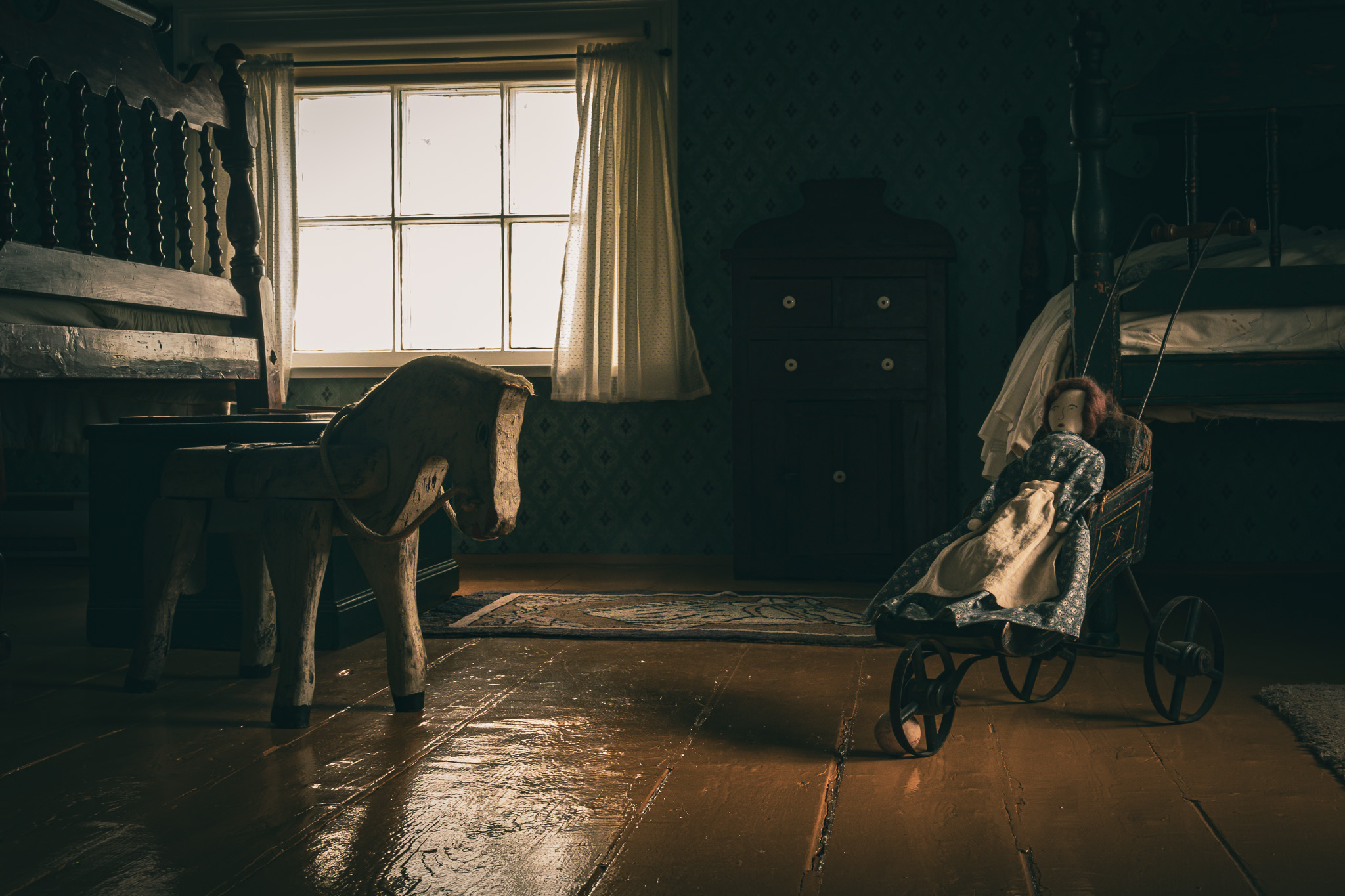 An darkly lit antique child's room in the Bonavista Lighthouse is perfectly preserved. A wooden horse stands next to a doll in an old stroller at the foot of two beds.