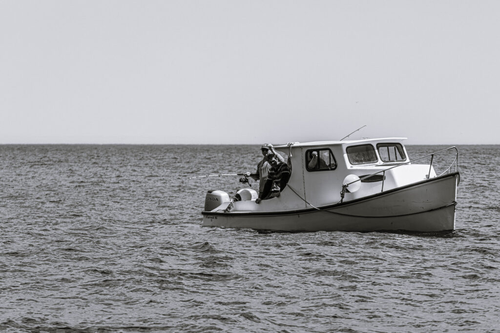A few locals with fishing rods stand on a small boat in the middle of the water. The image is black and white, and the people on the boat are shadowed.