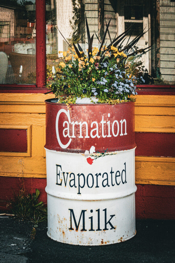 A large Carnation Evaporated Milk flower stand with rust spots sits in front of a yellow and red restaurant building. Yellow and blue flowers sit on top.