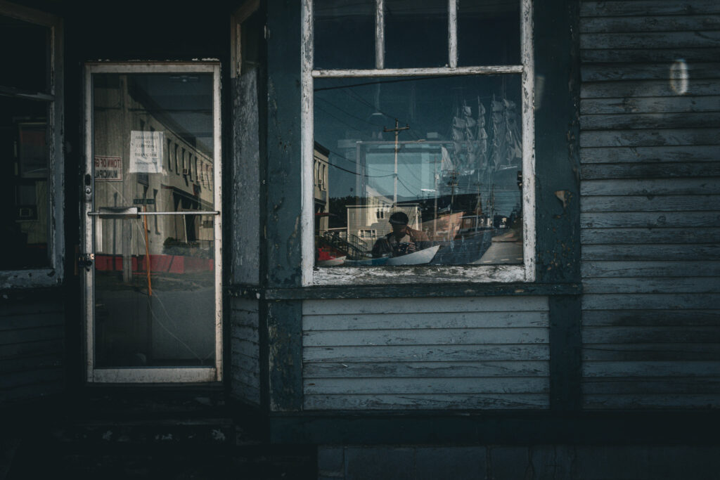 The storefront of a weathered, blue Newfoundland building. The store is dark inside, but a large ship model can be seen, as well as Rowan's reflection as he takes the photo.