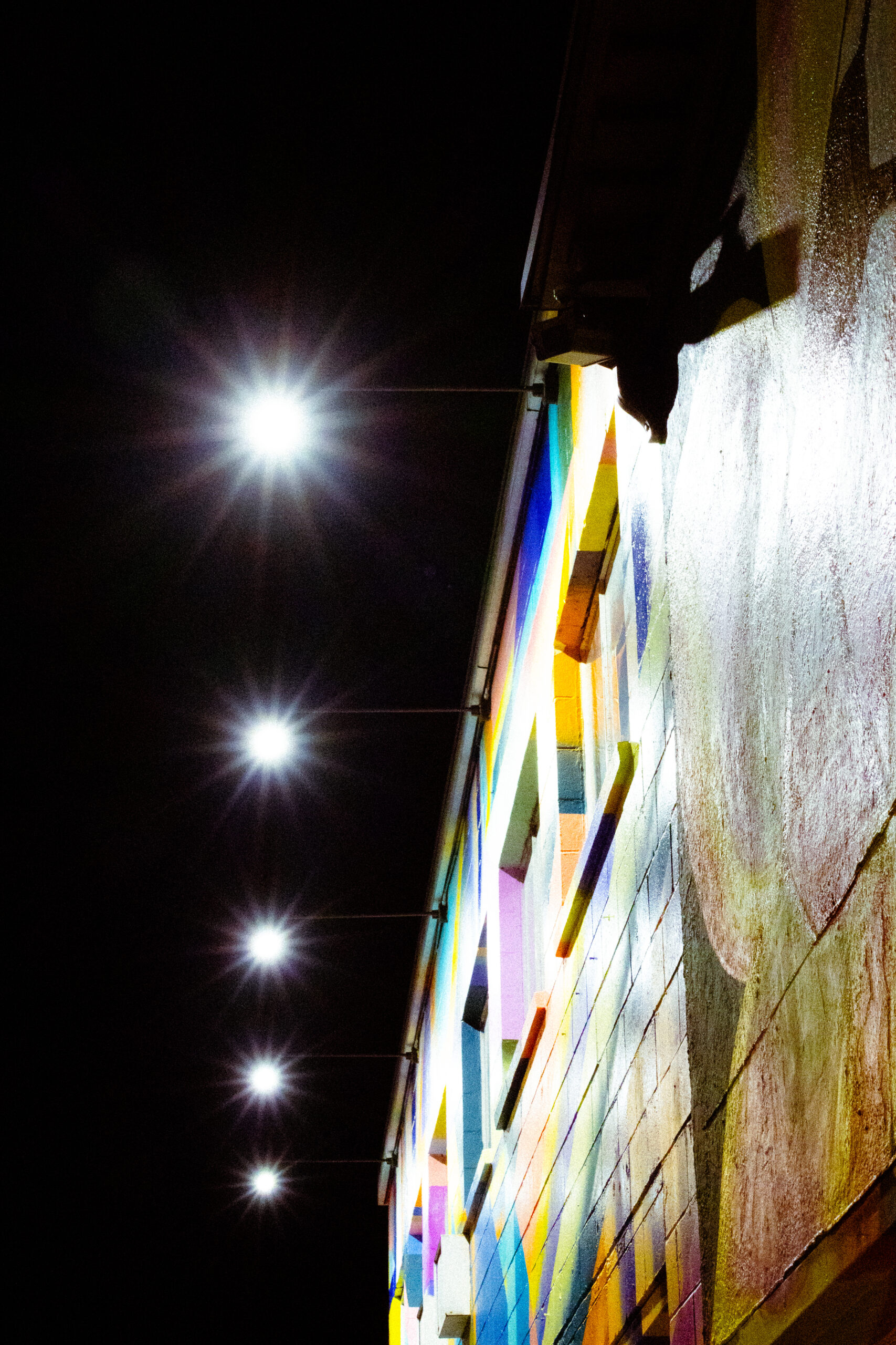 Lights shine down from the top of a building in a vertical row. They light the face of the building at night, which is painted in abstract shapes of different colours.